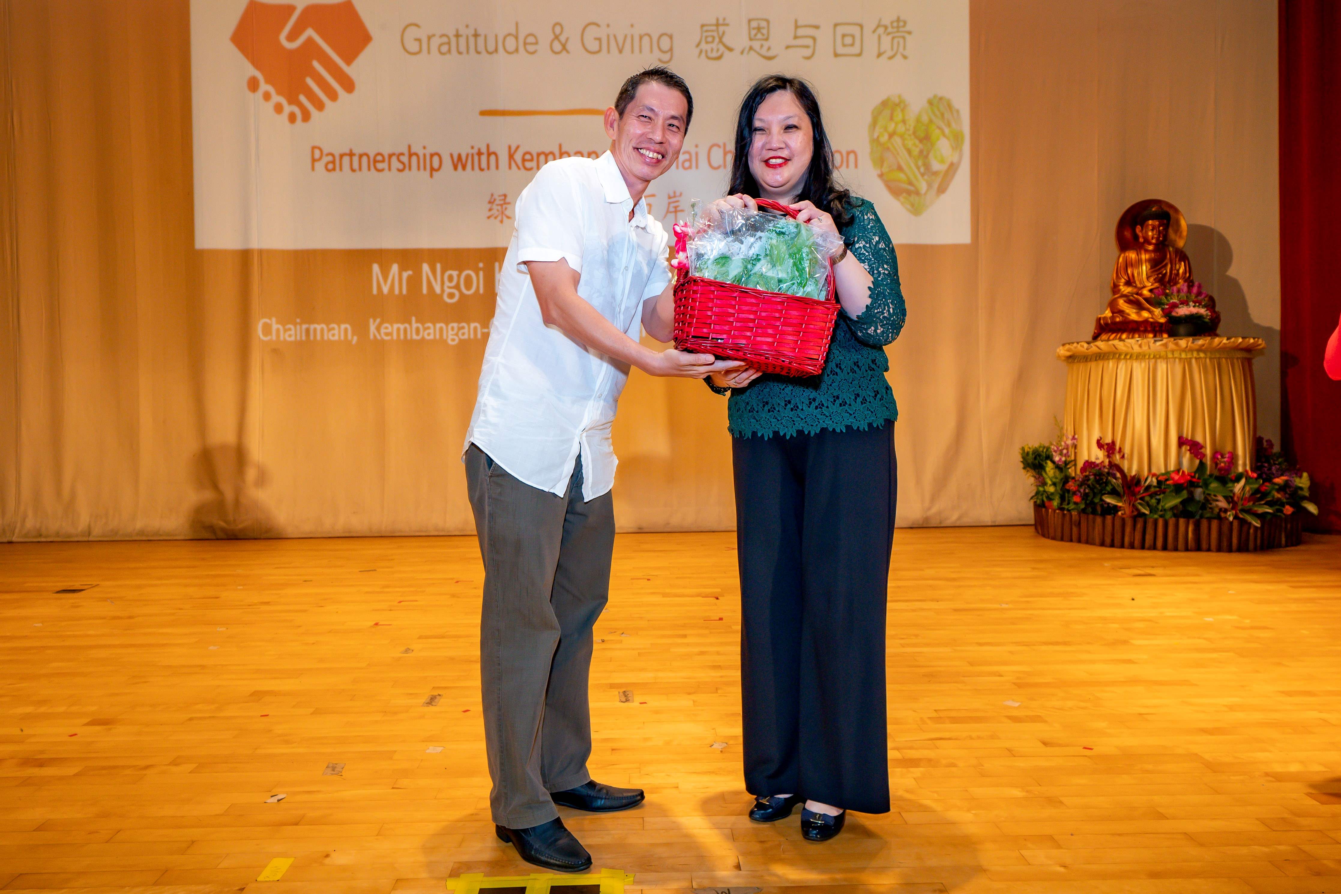 Two people smile on a stage holding a red basket with vegetables at a Gratitude and Giving event.